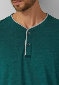 Türkisfarbenes Henley-Shirt aus strukturiertem Stoff mit grauem Kragen. Verfügt über zwei dunkle Knöpfe und ein kleines Logotag in der Nähe des Saums.