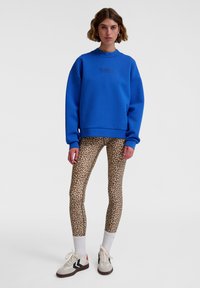 Blauer Sweatshirt mit Rundhalsausschnitt und Logo auf der Brust, kombiniert mit Leggings im Leopardenprint und Sneakers in neutralen Farbtönen. Weiße gerippte Bündchen sind sichtbar.