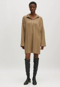 Langes braunes Kabelstrickkleid mit Reißverschluss vorne, gestaltet mit einem Kragen und weiten Ärmeln, kombiniert mit kniehohen schwarzen Stiefeln.