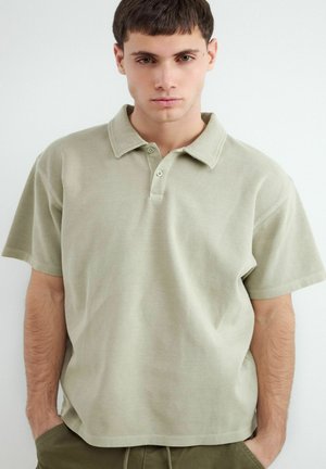 Junger Mann mit kurzen braunen Haaren, der ein hellbeiges Poloshirt und olivgrüne Hose trägt, steht mit den Händen in den Taschen vor weißem Hintergrund.