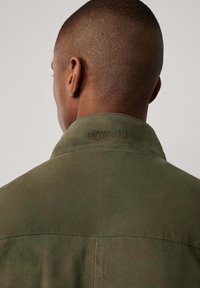 Chaqueta de color caqui con cuello de ante que presenta la etiqueta "FAÇONNABLE" grabada en el cuello, textura suave y diseño minimalista.