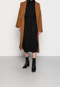 Cappotto in lana marrone con vestibilità sartoriale, indossato sopra un vestito nero a coste con cintura in vita. Abbinato a stivali neri alla caviglia.