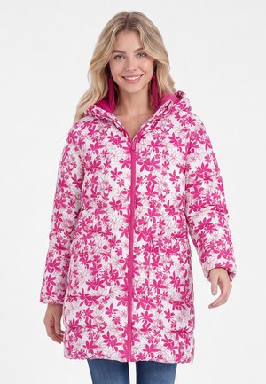 Lächelnde Frau, die eine weiße Steppjacke mit leuchtend pinkem Blumenmuster und Quastenohrringen trägt, steht vor einem schlichten Hintergrund.