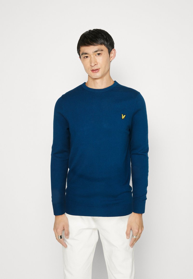 Maglione a maglia blu scuro con scollatura a girocollo, polsini e orlo a costine, caratterizzato da un piccolo logo giallo sul petto. Texture liscia.