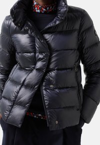 Marinblå puffjacka med blank yta, som har hög krage, horisontellt quiltad design och två framsydda fickor med dragkedja.