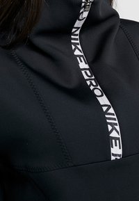 Zwarte Nike Pro sporttop met een hoge kraag, half-rits ontwerp en een opvallend wit logo langs de rits. Gladde textuur en een stijlvolle pasvorm.