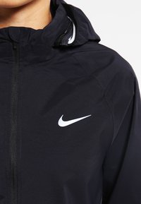 Personne portant une veste à capuche noire avec une fermeture éclair à l'avant et un logo blanc Nike Swoosh sur la poitrine.
