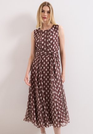Vrouw draagt een mouwloze bruine midi-jurk met grote witte stippen en een stoffen riem gestrikt in de taille.