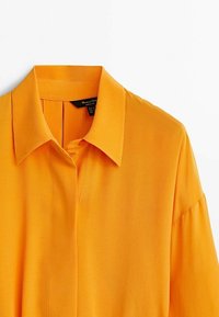 Chemise orange à manches longues avec un col pointu et une patte de boutonnage dissimulée, présentée sur un fond blanc.