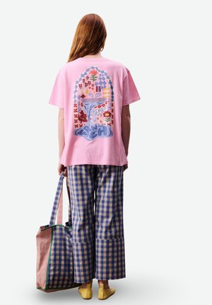 Personne aux longs cheveux roux portant un t-shirt rose à motif graphique, un pantalon à carreaux bleu et beige, des chaussures jaunes, tenant un grand sac fourre-tout à carreaux.