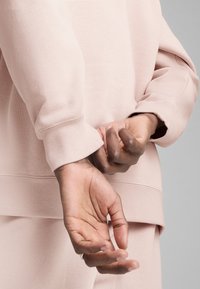 Lyserød sweatshirts med en afslappet pasform; har ribbede manchetter og en lige søm. Hænderne er placeret bag ryggen og holder let om hinanden.