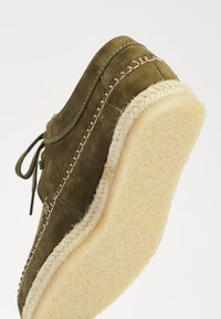 Chaussure en daim verte avec une forme arrondie, semelle en caoutchouc beige et coutures décoratives le long du bord. Les lacets sont visibles sur le dessus.