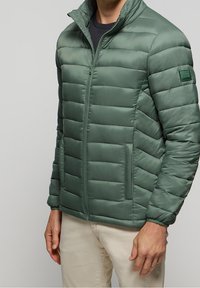 Zelená prošívaná péřová bunda se zapínáním na zip vpředu, vysokým límcem, dvěma bočními kapsami a nášivkou s logem na levém rukávu. Hladká textura.