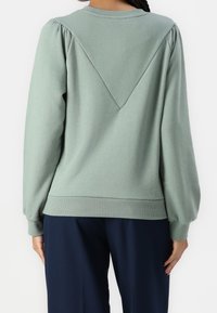 Sudadera verde con mangas abullonadas, que presenta un diseño en forma de V en la parte trasera, puños acanalados y un dobladillo con banda. Tela suave y texturizada.