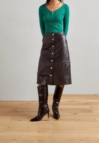 Haut vert à manches longues côtelé avec fermeture éclair, jupe midi en cuir noir avec boutons sur le devant, et bottes noires à talons hauts brillantes.