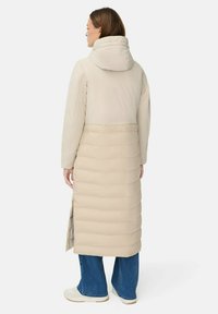 Kvinde iført en beige quilted lang frakke med hætte, blå vide jeans og hvide sneakers, stående med ryggen til på en hvid baggrund.
