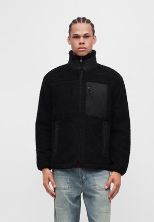 Jeune homme aux cheveux tressés portant une veste polaire noire zippée et un jean bleu clair usé, debout devant un fond uni.