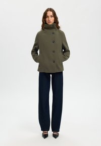 Olivgrüner Wollmischungs-Mantel mit hohem Kragen, fünf schwarzen Knöpfen und seitlichen Taschen. Kombiniert mit dunkelblauen weit geschnittenen Jeans und spitzen schwarzen Schuhen.