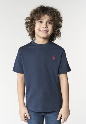 Bambino sorridente con capelli ricci che indossa una maglietta blu navy con logo rosso e jeans blu, in piedi contro uno sfondo chiaro e uniforme.