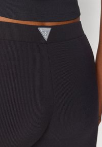 Czarne prążkowane legginsy z trójkątną, srebrną naszywką logo na pasku, podkreślające dopasowany krój i teksturowaną powierzchnię.