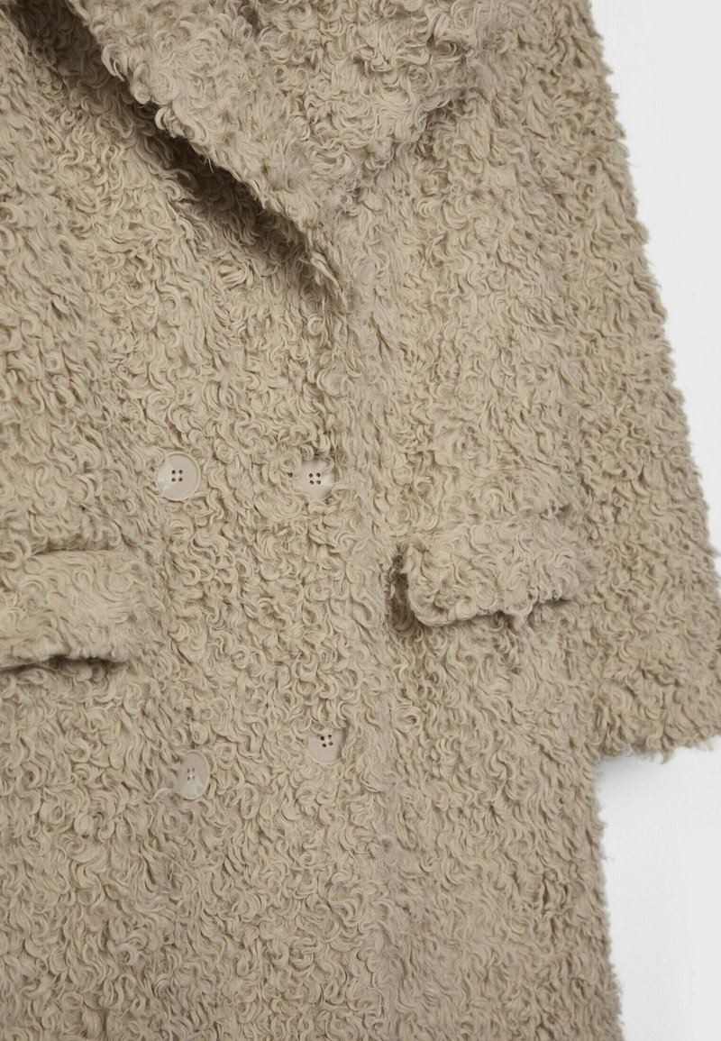 Beżowy, teksturowany płaszcz wykonany z kręconego materiału, z kołnierzem w kształcie klap, podwójnym zapięciem na guziki oraz bocznymi kieszeniami.