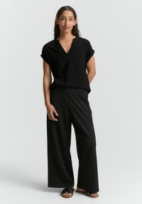 Schwarzes, locker sitzendes Oberteil mit V-Ausschnitt und aufgerollten Ärmeln, kombiniert mit schwarzen, weiten Hosen und flachen schwarzen Sandalen. Glatte Stofftextur.