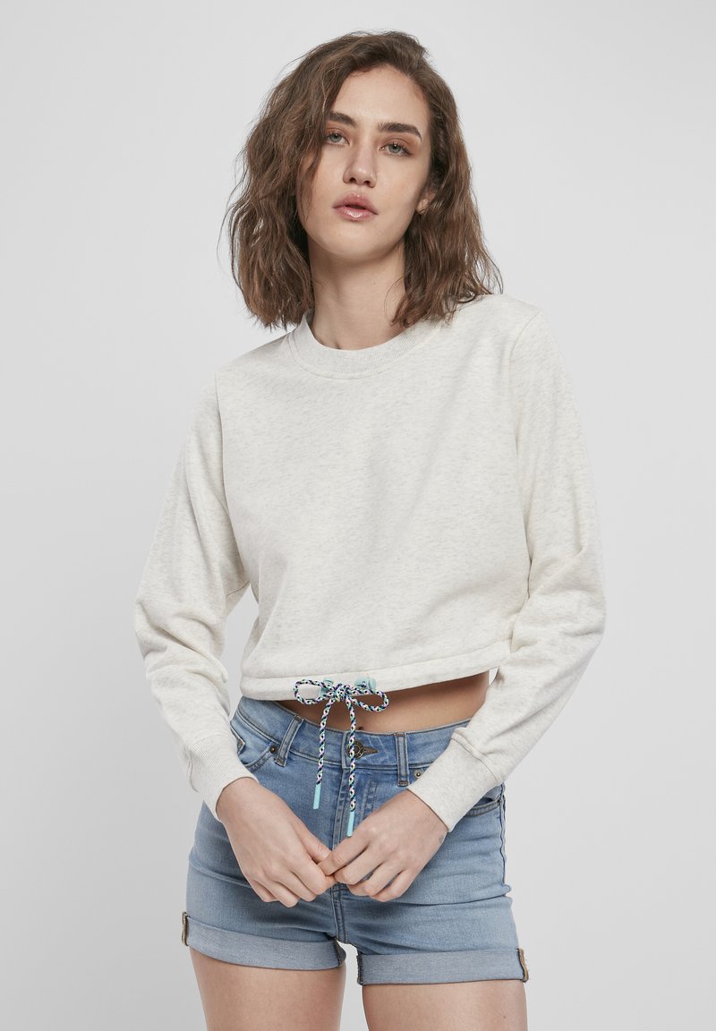 Kremfarget kort sweatshirt med lange ermer, med en snor i nederkant med et flerfarget mønster. Kombinert med lyse denimshorts.