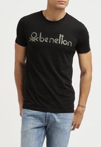 Černé bavlněné tričko s opotřebovaným zeleným logem „benetton“ napsaným přes přední část. Krátké rukávy a střih s kulatým výstřihem.