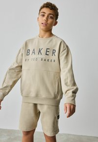 Sudadera beige con "BAKER" impreso en letras oscuras, con un bolsillo frontal; combinada con pantalones cortos a juego que tienen un bolsillo lateral.