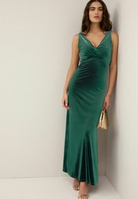 Robe en velours vert émeraude, longueur maxi, avec un décolleté en V, un corsage ajusté, une jupe évasée, et accompagnée d'un sac à main doré et de sandales à lanières.