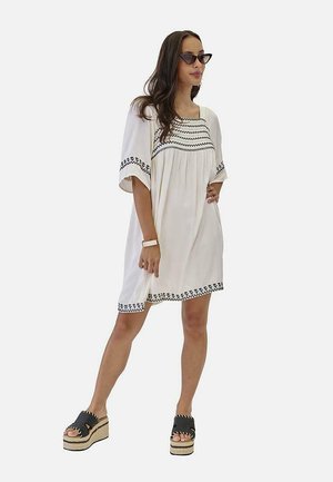 Mujer con vestido blanco hasta la rodilla con bordados negros en las mangas, el pecho y el dobladillo, sandalias de plataforma negras, gafas de sol y una pulsera.