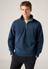 Φούτερ από μάλλινο ύφασμα σε ναυτικό μπλε με φερμουάρ στο λαιμό μέχρι τη μέση, ριμπ μανσέτες και τελείωμα. Διακριτική κεντημένη λογότυπη στο στήθος. Μαλακή υφή.