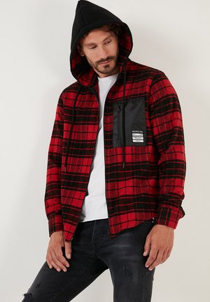 Cămașă din flanel în carouri roșii și negre, cu un glugă neagră, având un buzunar pe piept cu fermoar și șnururi de ajustare, purtată peste un tricou alb.