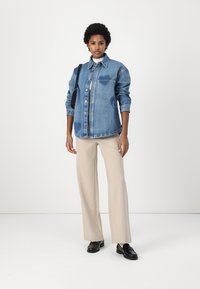 Lys blå denimskjortejakke med to brystlommer og knapper, kombinert med beige flagrende bukser og svarte sko. Enkelt design og passform.
