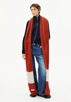 Une jeune personne se tient debout, vêtue d'une longue écharpe rouille et blanche, d'une veste zippée bleu marine sur un t-shirt noir, de jeans bleus et de chaussures noires à franges, sur un fond blanc.