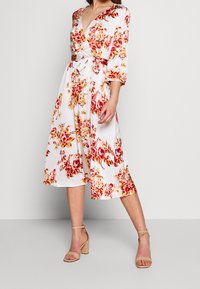 Robe portefeuille florale en tissu blanc à roses rouges et jaunes. Présente un décolleté en V, des manches trois-quarts et une taille ceinturée. Comprend des sandales à talons blocs beige.
