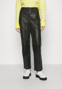 Person klädd i svarta läderbyxor, vita kraftiga stövlar och en gul kabelstickad tröja, stående mot en enfärgad bakgrund.