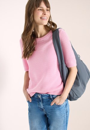 Femme portant un haut rose en maille à manches courtes et un jean bleu, souriante, avec un sac cabas gris sur l'épaule et les mains dans les poches.