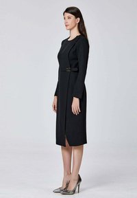 Robe noire à manches longues avec un ourlet fendu et un détail de boucle à la taille. Texture lisse, longueur genou, associée à des chaussures à talons métalliques.