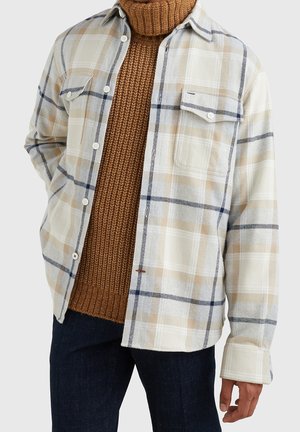 Homme portant une chemise à carreaux beige et blanche boutonnée sur un pull à col roulé marron tricoté et un pantalon foncé sur un fond uni.