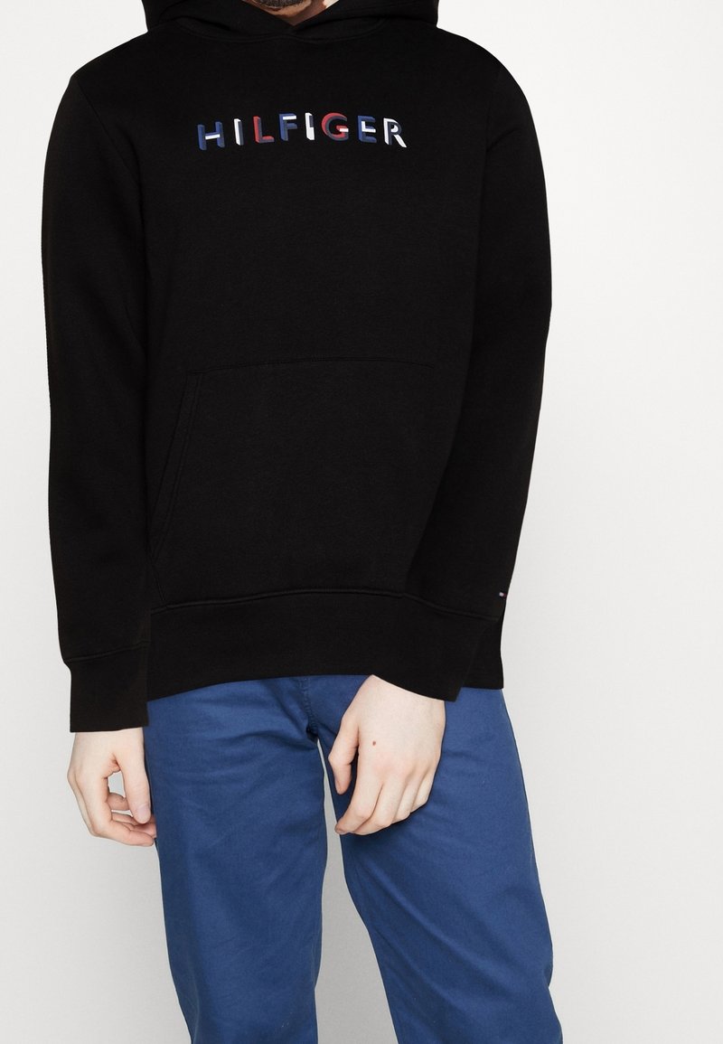 Hoodie noir avec "HILFIGER" en lettres multicolores sur la poitrine, porté avec un pantalon bleu, mains détendues le long du corps sur un fond uni.