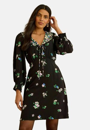 Frau mit langem dunklem Haar, die ein schwarzes Blumenkleid mit gepufften langen Ärmeln und einem gebundenen V-Ausschnitt trägt und mit einer Hand nahe am Ohr posiert.