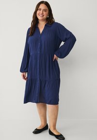 Vestido azul marino de botones con mangas largas, diseño en capas y una textura suave. Lleva zapatos planos negros y posa con las manos en las caderas.