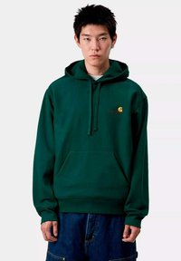 Sudadera con capucha verde y bolsillo delantero, con un pequeño logo naranja en el pecho. Está hecha de una tela suave y tiene un corte holgado.