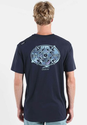 Homme aux cheveux blonds courts portant un T-shirt bleu marine avec un motif floral bleu clair et les mots "STAY ALIVE" et "OXBOW" au dos.