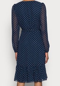 Laivansinisellä pilkkumekolla on pitkät läpikuultavat hihat, istuva vyötärö ja röyhelöhelma. Mekko on valmistettu kevyestä kankaasta, jossa on valkoisia pilkkuja.