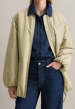 Personne portant une veste matelassée beige sur une chemise en denim bleu foncé et un jean bleu foncé taille haute, avec une main dans la poche.