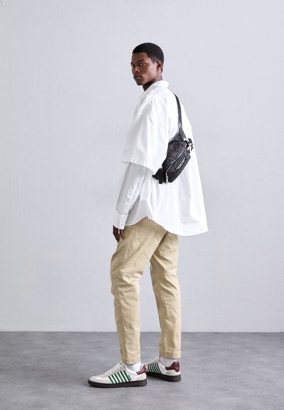 Black crossbody bag, oversized white shirt, beige pants, and green-striped white sneakers. Smooth textures, casual fit, minimal design.