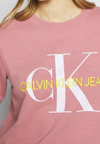 Sudadera de cuello redondo rosa, hecha de tela suave, que presenta un gran logo blanco "CK" y "CALVIN KLEIN JEANS" bordado en amarillo.