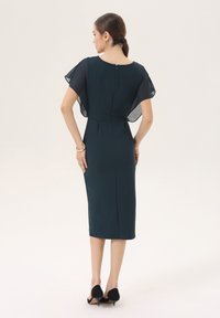 Robe à manches courtes en dentelle, de couleur vert foncé, arrivant au genou, avec une fermeture éclair au dos et une silhouette ajustée. Tissu texturé avec une finition lisse.